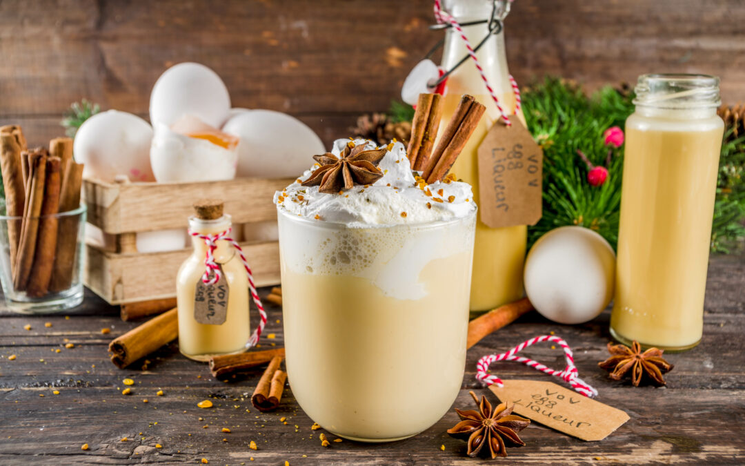 Ein Glas Bombardino mit Sahnehaube, serviert in einer Berghütte vor schneebedeckter Alpenkulisse.