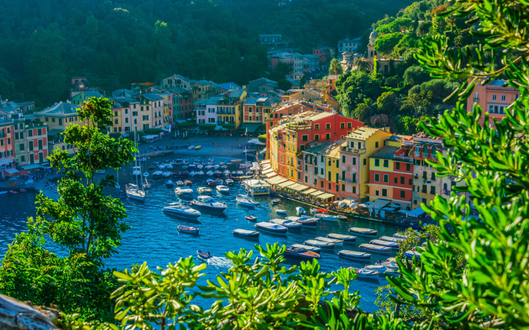 Panoramablick auf Portofino mit dem Hafen, bunten Häusern und dem Leuchtturm im Hintergrund, umgeben von grünen Hügeln und dem klaren blauen Meer."